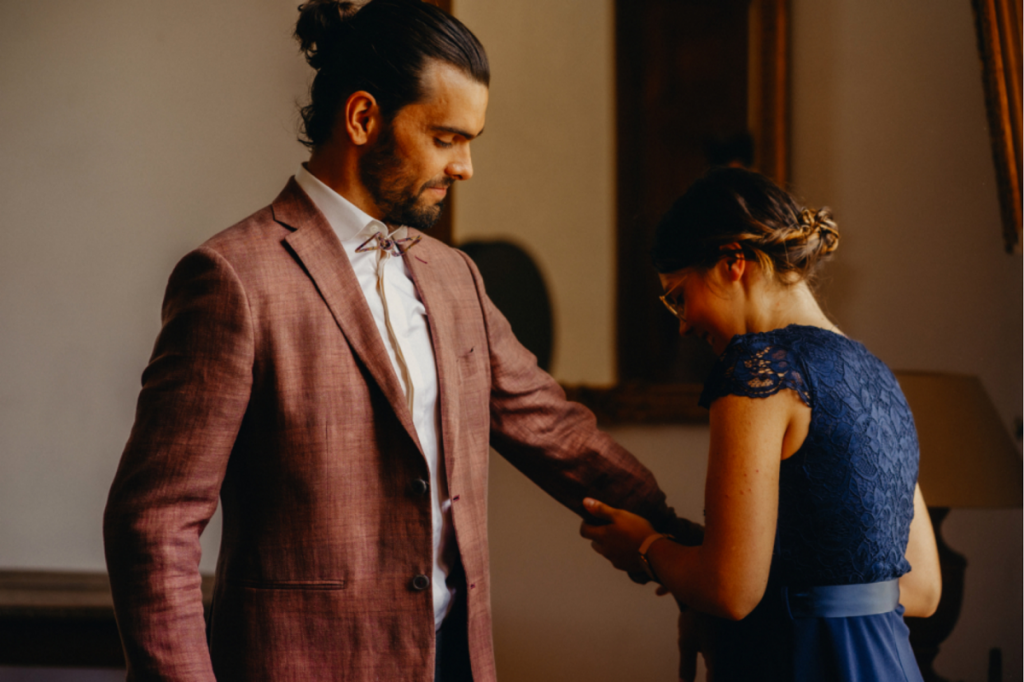 Comment choisir son costume mariage : la wedding planner ajuste la veste vieux rose sur le marié