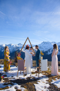 Mariage à la montagne cérémonie laïque les mariés au centre avec derrière eux l'officiante l'arche triangulaire et les montagnes. De part et d'autre les témoins